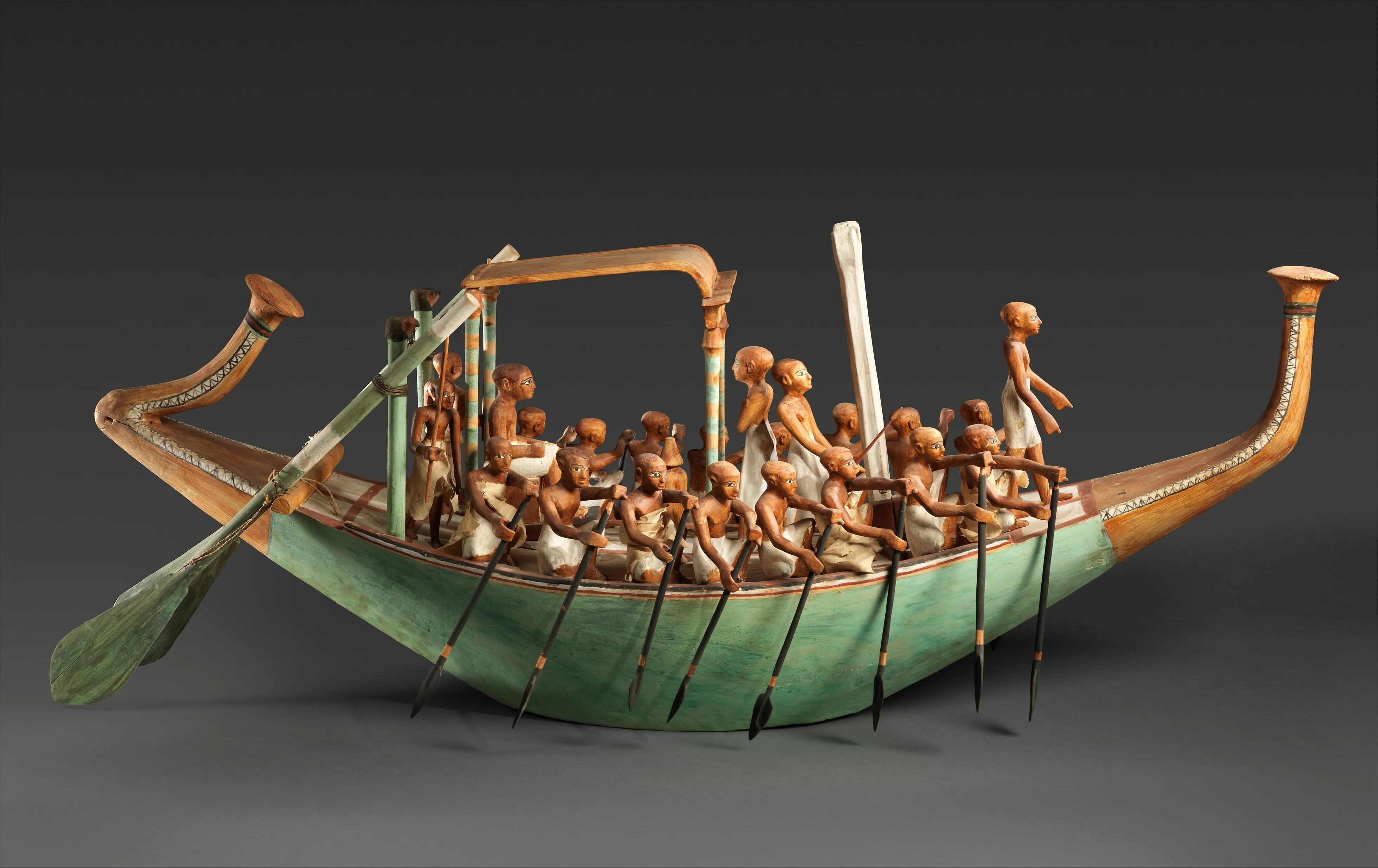 Side view of an ancient Egyptian wooden model with sixteen kneeling paddlers along a green hull and two long steering oars at the raised stern.